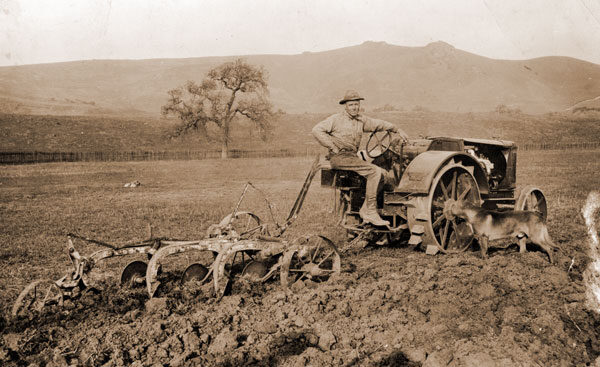 Historical sepia-toned photo of a man plowing a field with an early tractor
