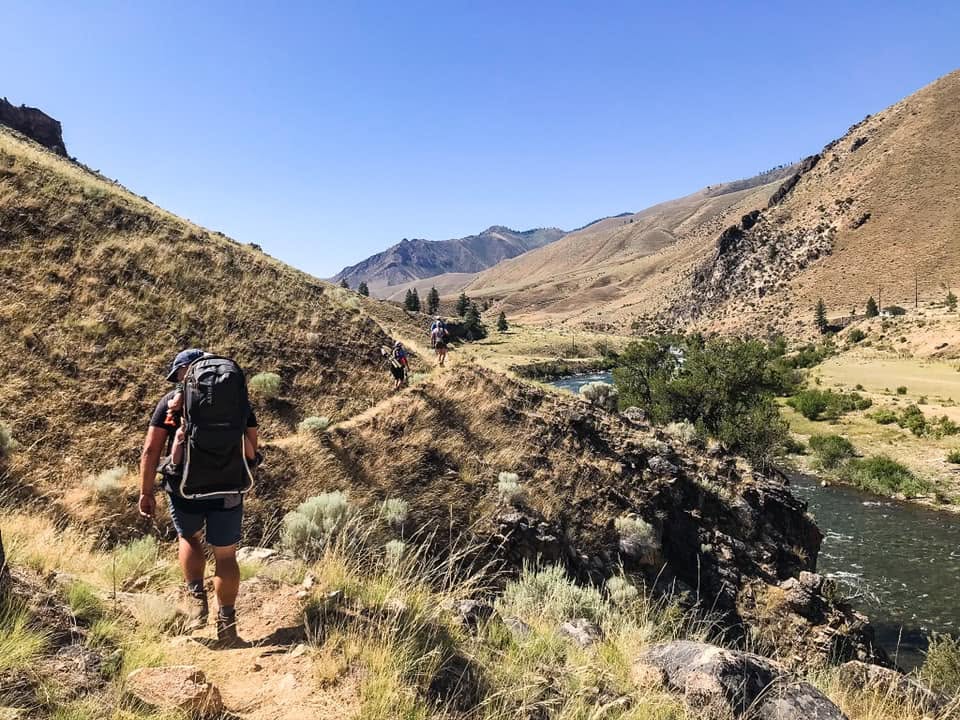 Hikers with backpacks descending a canyon trail alongside a river, with distant hikers ahead