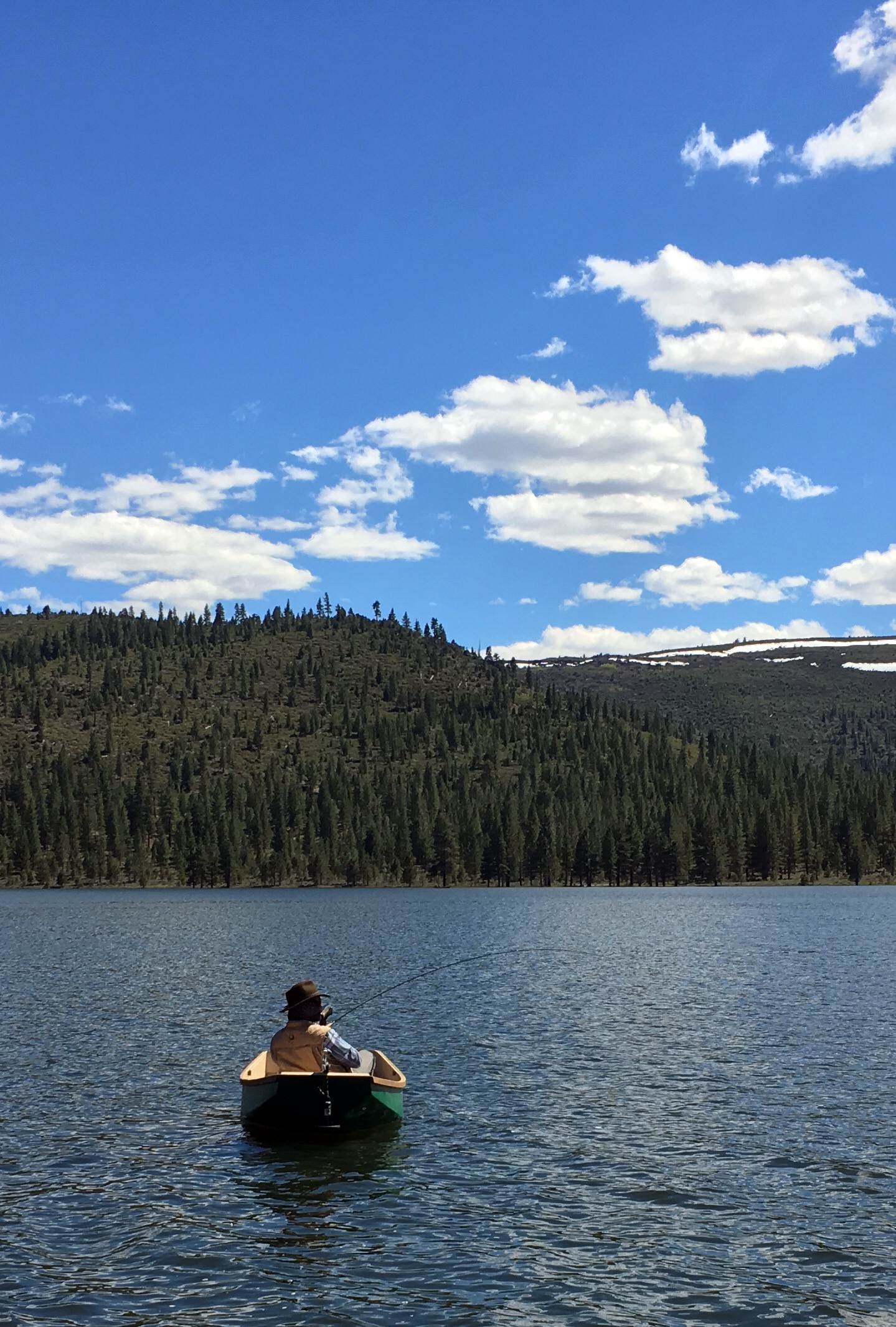 A man fly fishing from a canoe on Round Valley Reservoir on a sunny day, forested hills in the background